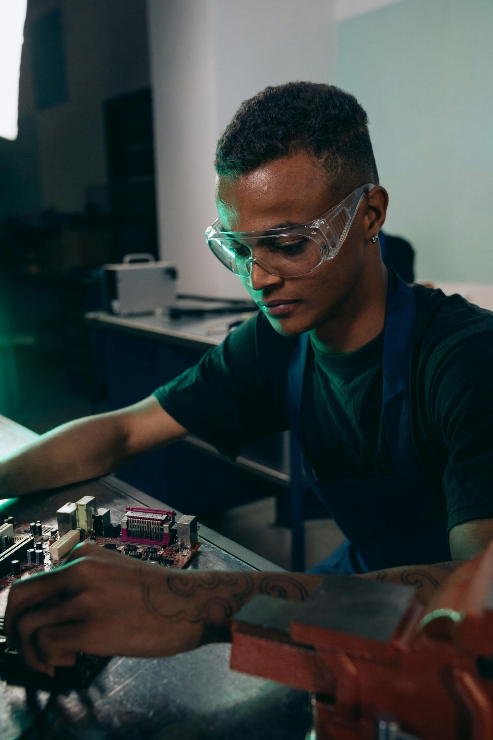 Focused young engineer working on an electronic circuit board in a workshop setting.